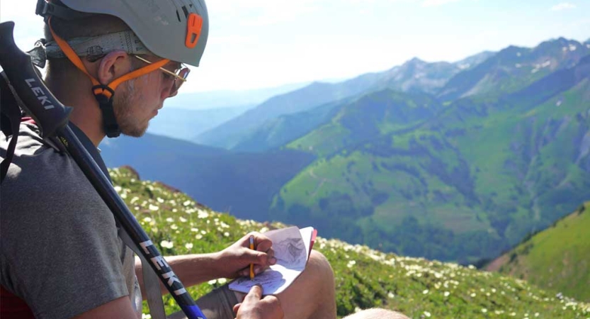 A person wearing a helmet sits at a high elevation and journals. Beyond and below them is a vast mountainous landscape. 